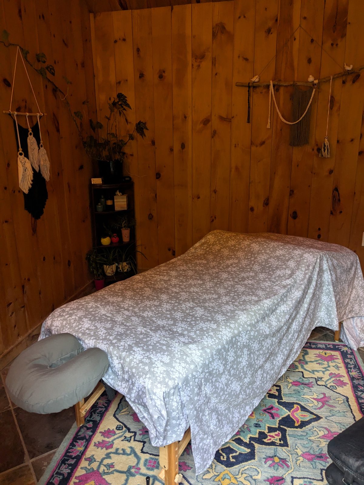 The treatment room: a massage table made up with a floral sheet and face
                cradle, against pine-panelled walls, with a patterned rug, plants and a
                small shelf of oils.